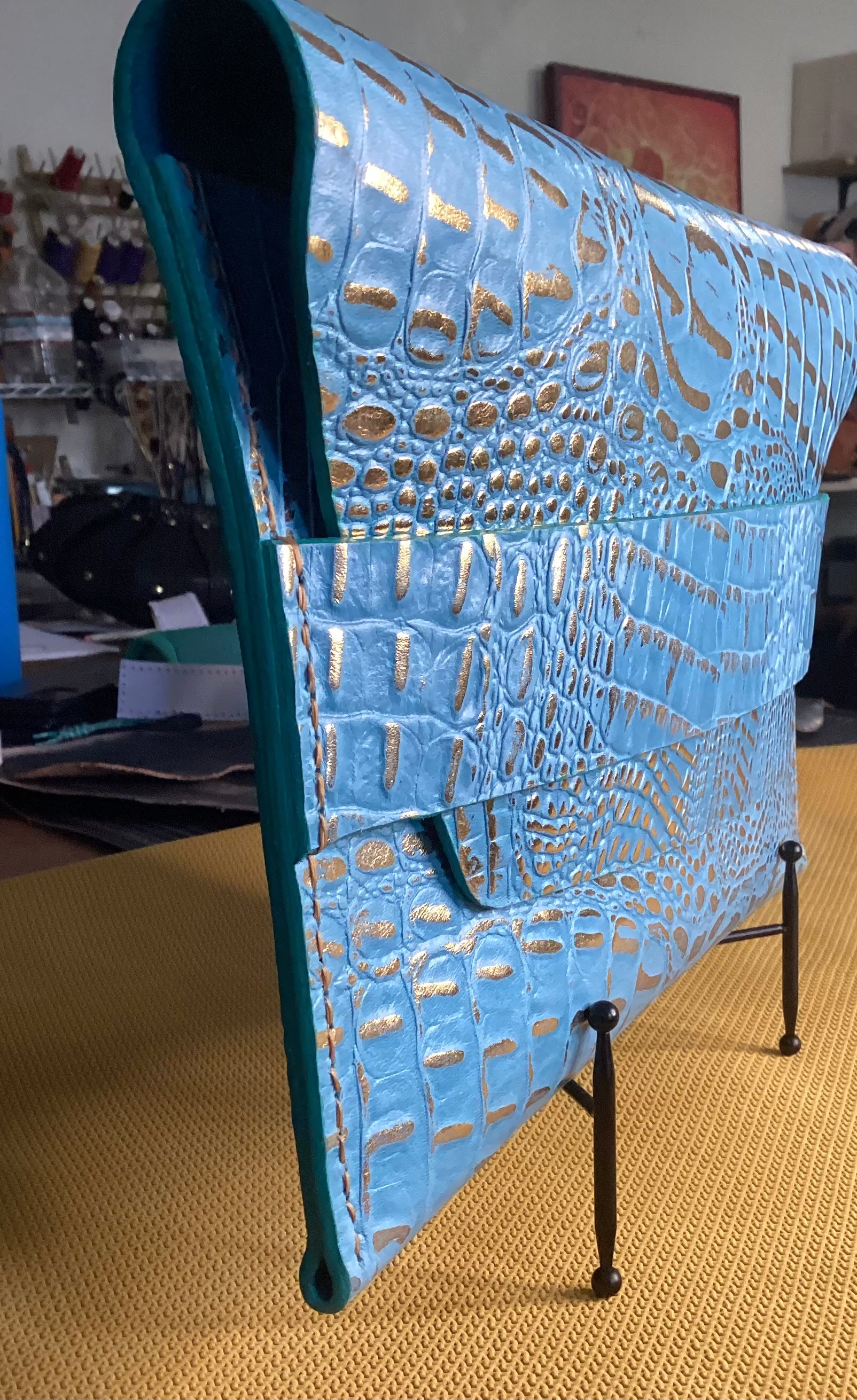 “Turquoise leather envelope clutch with polished gold accents, angled view.”